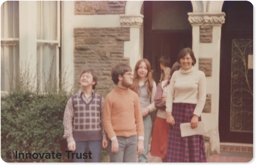 This photograph shows a group of people standing outside a house which they shared. They are students and people with learning disabilities. The image is from the 1970s.