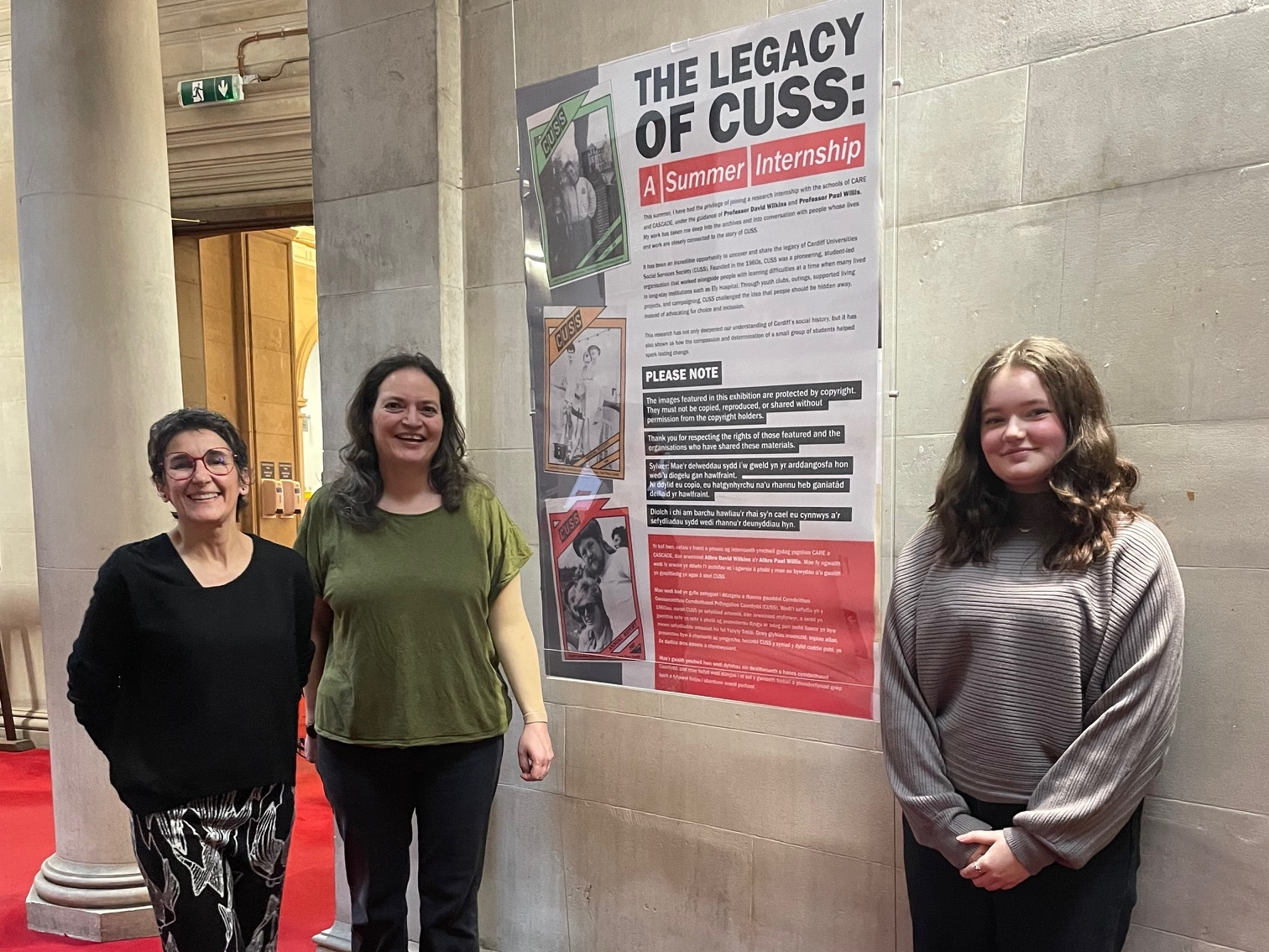 Three white women stand in front of a large poster with the title "The Legacy of Cuss: A Summer Internship".