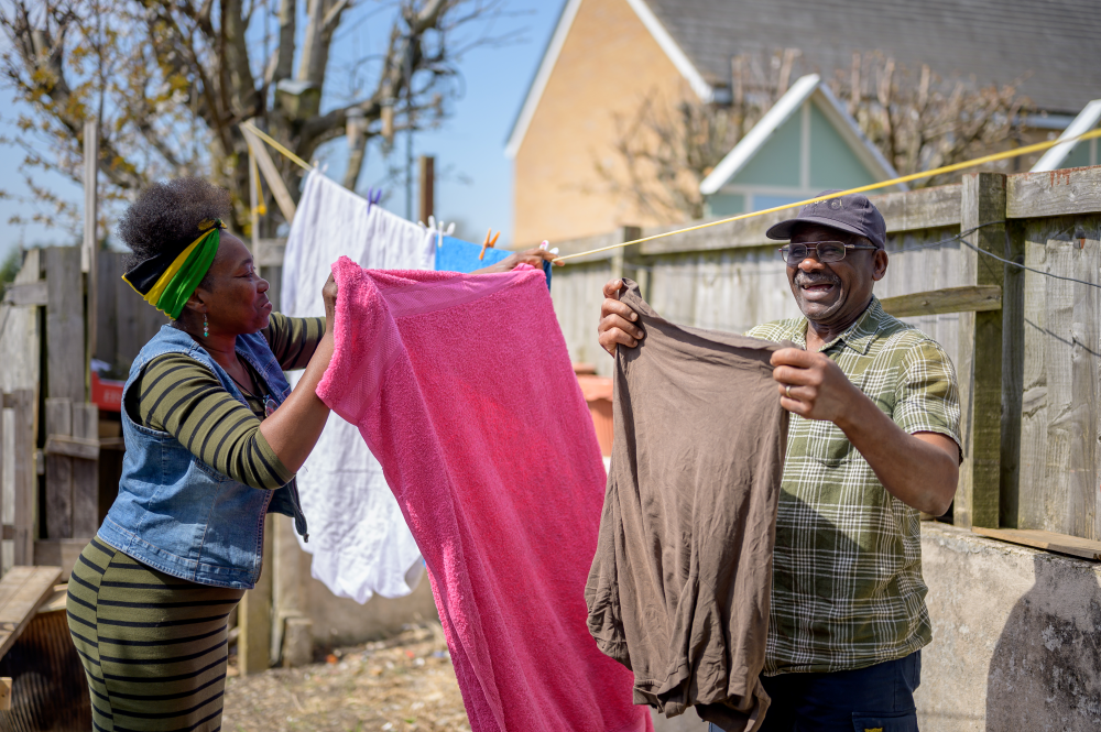 Two people hang up washing in the back garden on a sunny day. There is an older man and a younger woman and they are both black.